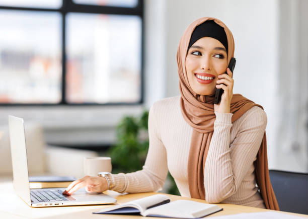 istock Friendly smiling muslim woman  smiling and making call via smartphone while working remotely from home while sitting at desk and talking to coworkers 1494066466