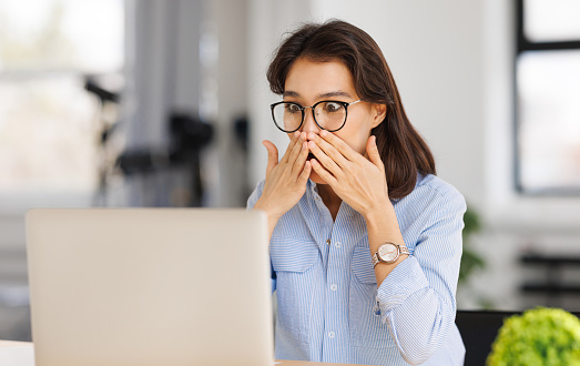 Shocked   woman freelancer looks in amazement at laptop screen and covers her mouth with hands in surprise while sitting at desk in home office