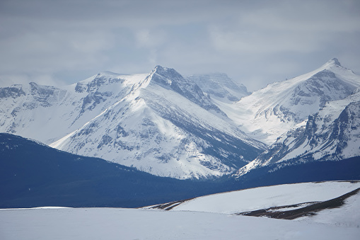 Snow-covered mountain slopes fold one into the other and rise into frozen peaks.  A frosty hillside is darker and marks the space between the snowy plains and frozen mountains. Wispy clouds fill the sky above continuing the powder blue pallet of the scene on the Eastern edge of Glacier National Park, Montana.