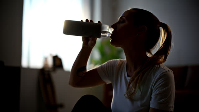 SLOW MOTION Silhouetted tired,fit young woman with ponytail drinking water from water bottle after home workout on living room floor. Dedication,exercise,healthy lifestyle. Shot in 8K Resolution.