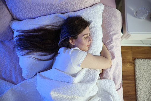 Serene beautiful woman sleeping with hand under cheek on soft pillow, young female wearing white pajamas resting under warm blanket in comfortable bed in bedroom or in hotel, enjoying sweet dreams