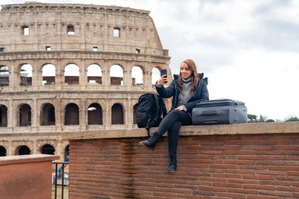 happy young latin woman tourist with luggage and warm clothes taking selfie on smartphone in front of historic building colosseum in rome, italy against cloudy sky - colosseum visit stockfoto's en -beelden