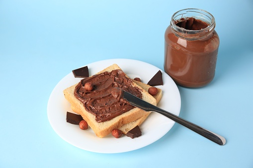 Tasty toast with chocolate paste and nuts near jar of cream on light blue background