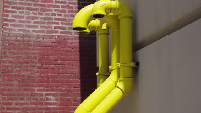 Three strange bright yellow pipes just out from the side of a white cement building in front of an old red brick wall