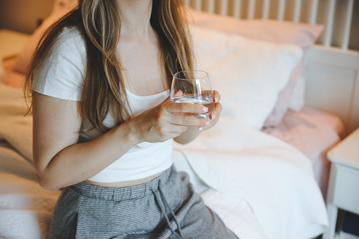A glass of water next to a girl sleeping in bed. Happy morning. Woman in pyjamas. Healthy lifestyle, wellness. Proper nutrition. Drinking water. Morning with water. Sunlight on linens.