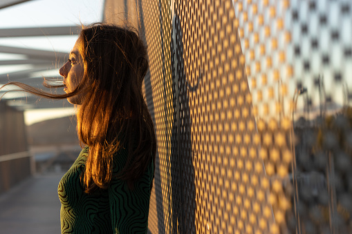 Side view of a young woman standing on a pedestrian bridge against steel mesh looking to sunset.