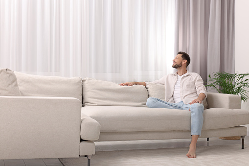 Happy man on resting sofa near window with beautiful curtains in living room. Space for text