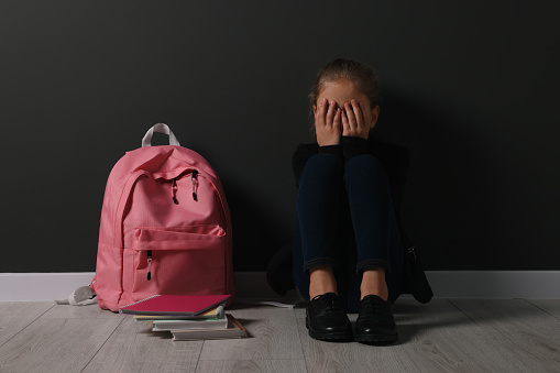 Upset girl with backpack sitting on floor near black wall. School bullying