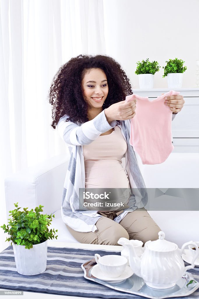 Waiting for a newborn child A beautiful, smiling pregnant young adult woman sitting on sofa in living room and looking at baby clothes. Pregnant Stock Photo