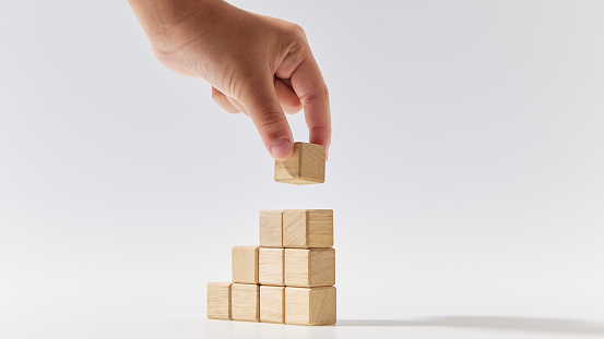 Close-up hand putting wood cubes stacked as stair step shapes mock-up for creating symbols or logos, business growth, and management concept on white background