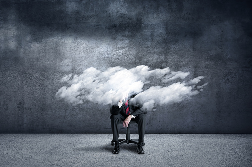 A businessman places his head in hands as he sits in an office chair under a cloud of stress. His head is obscured by the clouds.