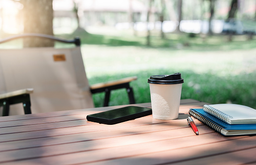 Cup of coffe and smartphone, note book on wooden table with lawn chair. coffee break and relax after work in parks. No people, Blurred  background