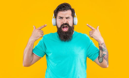 studio shot of displeased annoyed man by loud music. annoyed man has loud music in headphones isolated on yellow. annoyed man listen loud music in headphones. annoyed man in headphones with loud music