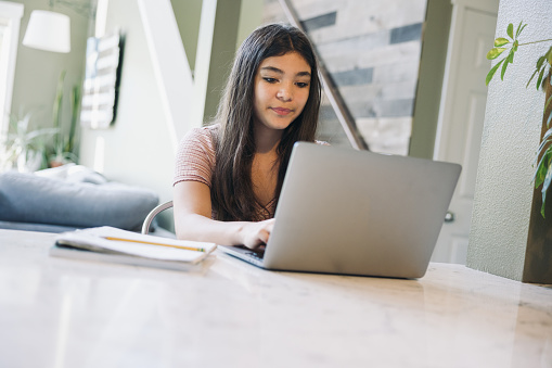 A Hispanic young  woman uses a laptop and textbook to do school work at home.  Self discipline to study in free time.   She sits at a well lit kitchen island.