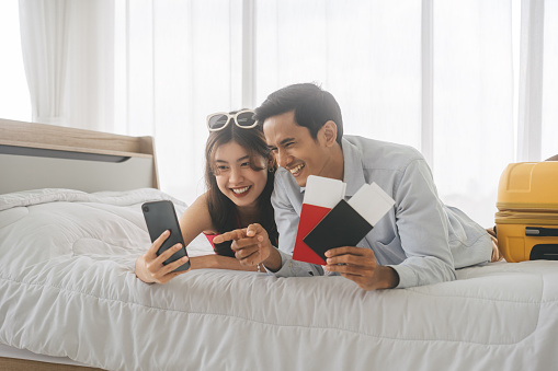 Happy smile young adult southeast asian couple using mobile phone for getting ready for holidays travel trip. Living at home laying on bed together. Curtain on background with day light