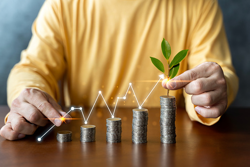 The green tree that is growing on coins increases in various forms, including the hands of business people that surround the pile of coins and the rising arrow graph. The concept of business growth.