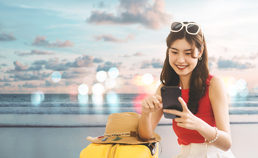Young adult southeast asian traveler woman with travel luggage. Using smartphone buy airplane ticket online cashless payment. Summer beach banner backgroud with copy space.