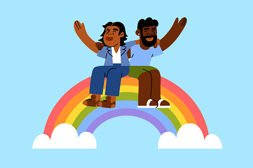 A black and brown gay male couple hold each other celebrating pride month sitting on a rainbow