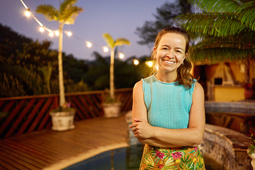 Smiling woman standing with her arms crossed outside on a patio illuminated with string lights on a summer evening