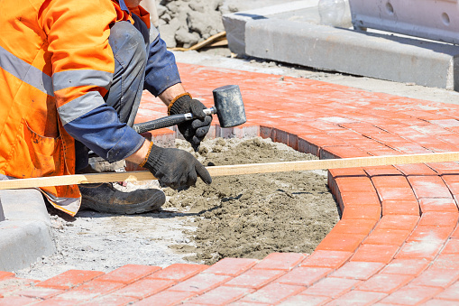 A bricklayer in work gloves and overalls carefully measures the size with a wooden plank and lays orange paving slabs on a footpath on a sunny day.
