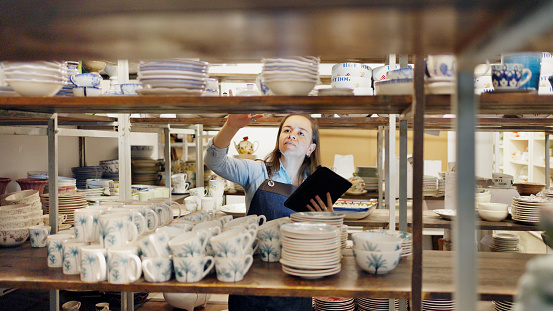 Female shop owner taking an inventory of items sitting on shelves in her ceramics store using a digital tablet