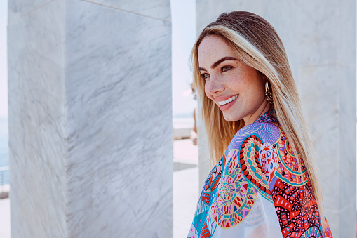 Side view portrait of a young beautiful blond latin american woman enjoys summer vacations. Female outdoors wearing colorful smock with marble column background.