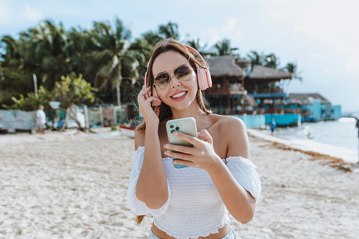 Relaxed Latin woman using mobile phone and wearing headphones listening to music at Caribbean Beach in Mexico Latin America, hispanic female