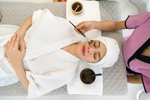 Young asian woman with nutrient facial dressed in bathrobe, getting facial care by beautician at spa salon, Top View. Face peeling mask, spa beauty treatment, skincare. Spa concept