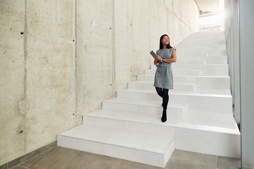 Happy Asian entrepreneur holding laptop while moving down the stairs in a hallway of an office building. Copy space.