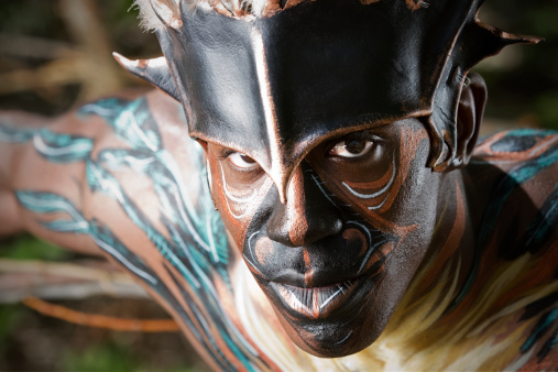 A handsome African American man with body paint and leather headdress looks intensely at the camera.
