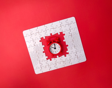 An alarm clock sitting atop a bright red jigsaw puzzle piece against a white background