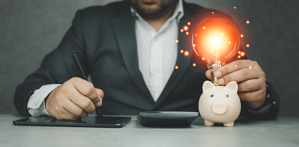 Businessman holding lightbulb on piggy bank with take note in digital tablet and calculator to calculate. Save energy and money with accounting finance. The idea of energy saving planning in the home.