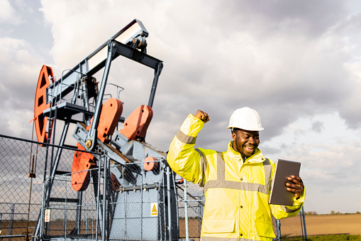 Oil field worker in protective work wear and helmet holding tablet by the oil rig.Portrait of an oil field worker standing in front of oil rigs drilling crude oil.