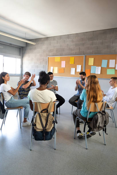 istock Multiracial high school students sitting in a circle clapping together celebrating achievement. Support group. Vertical. 1494236402