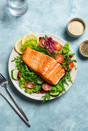 Grilled salmon fish fillet and fresh vegetable salad with tomato, lettuce, arugula. Healthy keto food - green salad and roasted salmon on blue table.