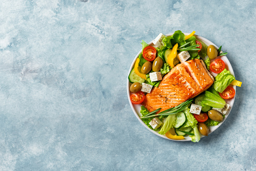 Grilled salmon fish fillet and fresh vegetable salad with tomato, olives, lettuce, feta cheese. Mediterranean food - greek salad and roasted salmon on blue background, copy space.