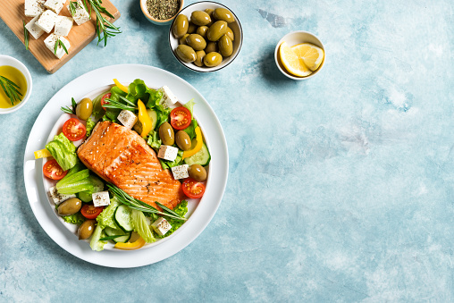 Grilled salmon fish fillet and fresh vegetable salad with tomato, olives, lettuce, feta cheese. Mediterranean keto food - greek salad and roasted salmon on blue background, copy space.