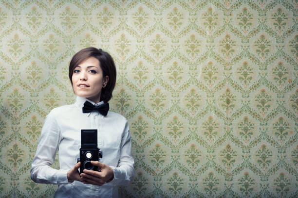 Short-haired woman, professional photographer, proudly grips a black twin-lens reflex camera. Dressed in white shirt, black bow tie, with dated but captivating wallpaper behind. Ready to teach darkroo