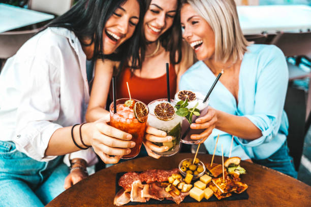 tres hermosas mujeres jóvenes bebiendo cócteles sentadas en la mesa del bar restaurante - amigas felices disfrutando de las vacaciones de verano al aire libre - concepto de estilo de vida con novias pasando el rato juntas - aperitivo bebida alcohólica fotografías e imágenes de stock