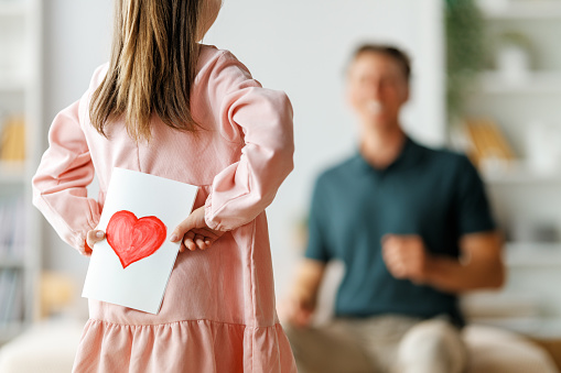 Child daughter is congratulating her father and giving him postcard. Dad and girl are smiling and hugging. Family holiday and togetherness.