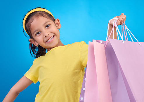 Girl portrait, shopping and gift bags from birthday party, event or celebration with a present in studio. Shop, bag and happy child looking at presents with isolated and blue background with a kid