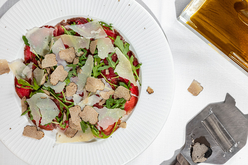 Italian dish. Veal carpaccio with ground black pepper and balsamic, arugula leaves, parmesan cheese and truffle on top. The food lies in a white, round ceramic plate, the plate stands on a table with a white tablecloth. Nearby is a glass of red wine on a high stem, a truffle grater and a truffle.