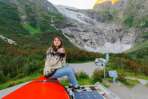 Young Caucasian woman in knitted sweater sitting on the rooftop of camper van on the background of glacier in Norway