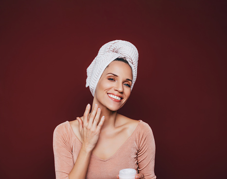 A portrait of a happy Caucasian woman with hair wrapped in a towel putting day cream on her neck.