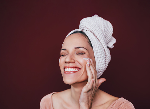 A portrait of a smiling Caucasian woman applying face cream on her cheeks.