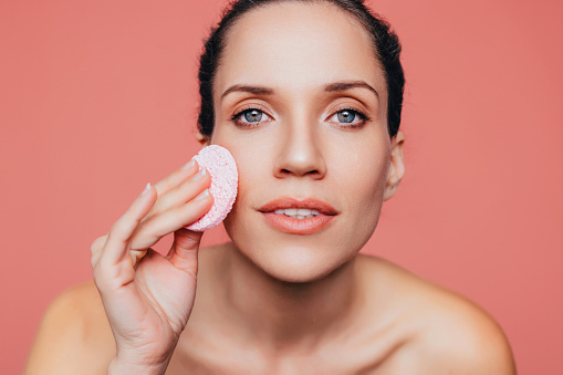 A portrait of a smiling Caucasian woman with blue eyes removing makeup with a sponge pad.