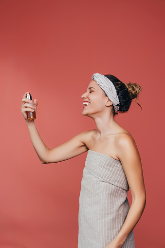 A happy woman with a hairband on her head applying spray moisturizer for face (coral color background).