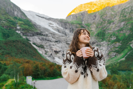 Young Caucasian woman in knitted sweater drinking coffee on the background of the glacier