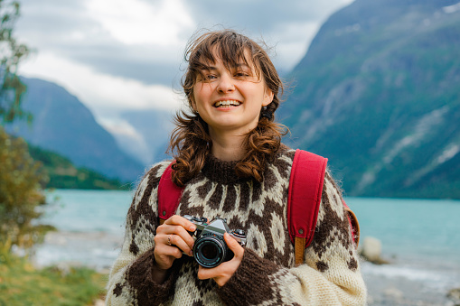 Young Caucasian woman in knitted sweater standing with vintage camera and photographing scenic views of the lake in Norway