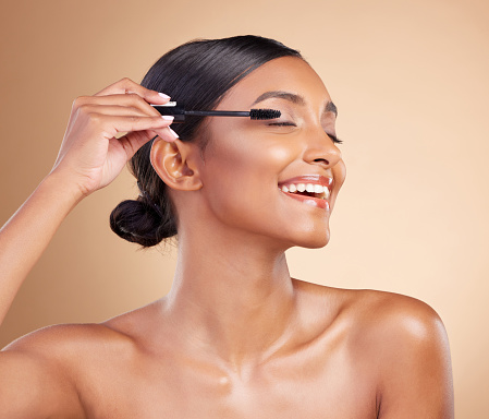 Makeup, happy and woman with mascara for eyelashes isolated on a studio background. Smile, cosmetology and Indian girl with a brush for applying cosmetics, pomade product or treatment to lashes